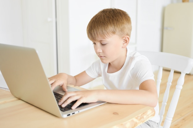 Portrait of handsome serious Caucasian boy of school age sitting at wooden table using laptop computer, keeping hands on keyboard. Education, leisure, people and modern electronic gadgets concept