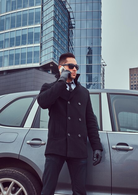 Portrait of a handsome serious male bodyguard in sunglasses talking on the phone near car outdoors.