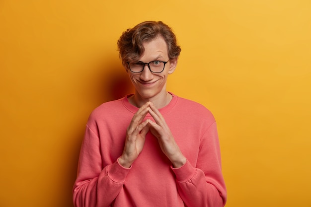 Portrait of handsome sly man steepls fingers, has evil plan, thinks about making surprise for girlfriend, has glad cunning expression, wears optical glasses and casual sweatshirt, poses indoor