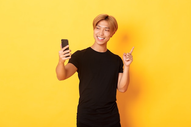 Portrait of handsome smiling asian man having video call on smartphone and pointing finger at something, standing yellow wall