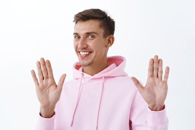 Portrait of handsome smiling blond man in pink hoodie, raising hands in surrender or sorry gesture, refuse or rejecting offer politely, standing white wall