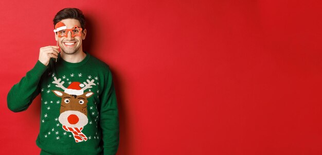 Портрет красивого улыбающегося человека в рождественском свитере и партийных очках, празднующего новый год и веселого, стоящего на красном фоне