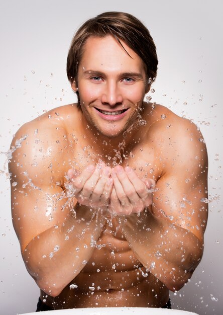 Portrait of a handsome smiling man washing his healthy face with water on gray wall.