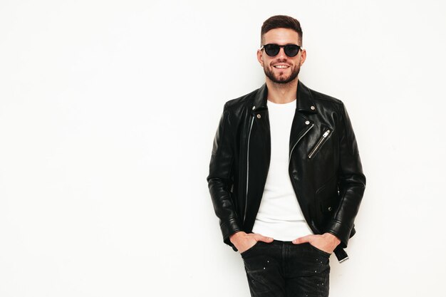 Portrait of handsome smiling model Sexy stylish man dressed in biker leather jacket and black jeans Fashion hipster male isolated on white in studio In sunglasses