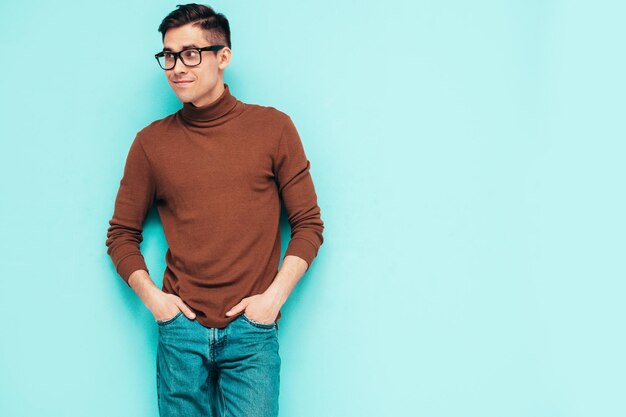 Free photo portrait of handsome smiling model sexy stylish man dressed in brown turtleneck sweater and jeans fashion hipster male posing near blue wall in studio isolated