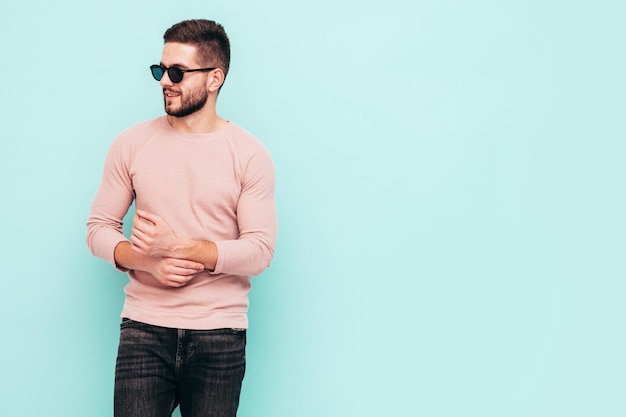 Portrait of handsome smiling model Sexy stylish man dressed in pink sweater and jeans Fashion hipster male posing near blue wall in studio In sunglasses Isolated