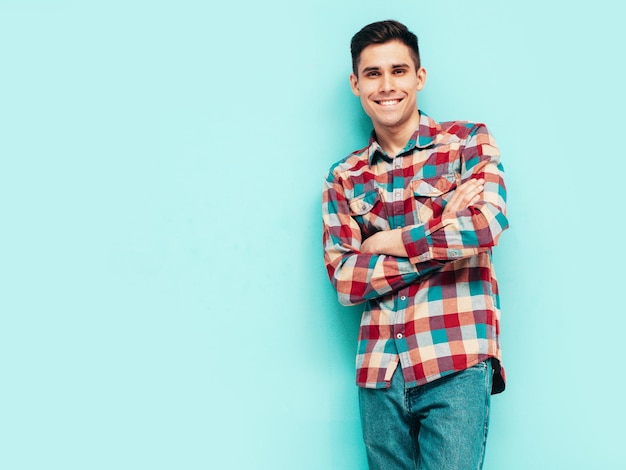 Portrait of handsome smiling model Sexy stylish man dressed in red checkered shirt and jeans Fashion hipster male posing near blue wall in studio Isolated Crossed arms