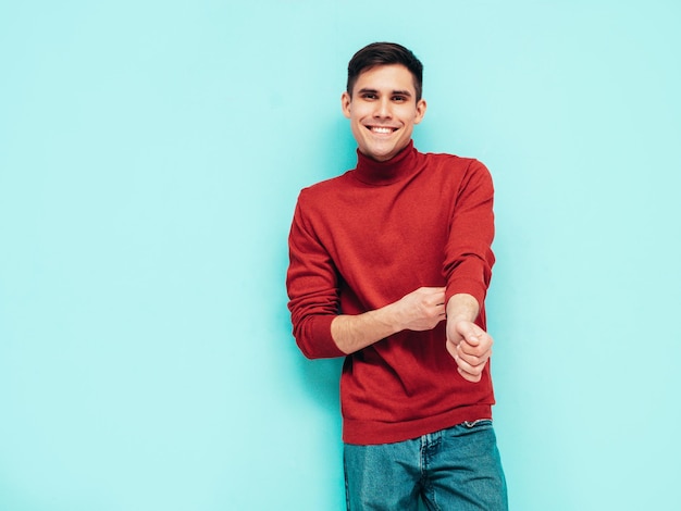 Portrait of handsome smiling model Sexy stylish man dressed in red turtleneck sweater and jeans Fashion hipster male posing near blue wall in studio Isolated