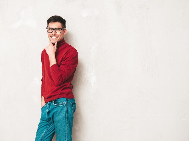 잘 생긴 웃는 모델의 초상화 빨간 터틀넥 스웨터와 청바지를 입고 섹시 한 세련 된 남자 절연 스튜디오에서 회색 벽 근처 포즈 패션 힙 스터 남성