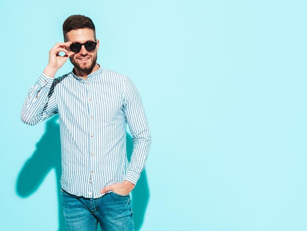 Portrait of handsome smiling model Sexy stylish man dressed in shirt and jeans Fashion hipster male posing near blue wall in studio Cheerful and happy