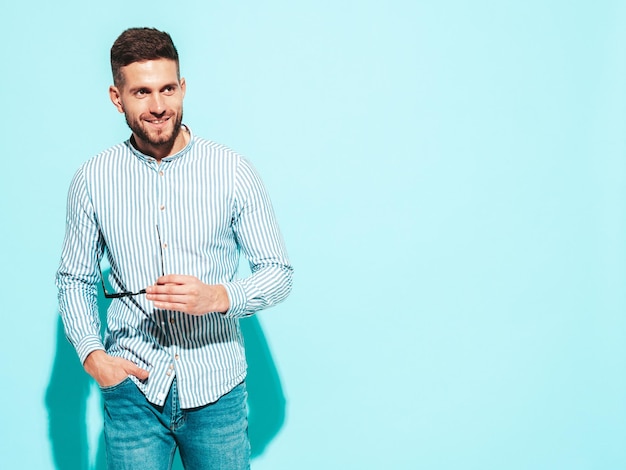 Portrait of handsome smiling model Sexy stylish man dressed in shirt and jeans Fashion hipster male posing near blue wall in studio Cheerful and happy