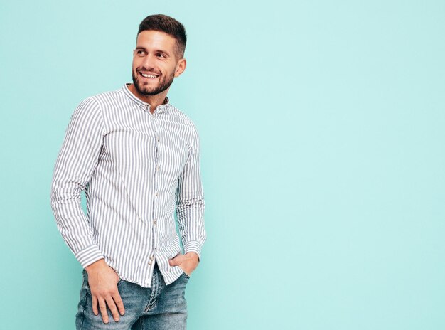 Portrait of handsome smiling model Sexy stylish man dressed in shirt and jeans Fashion hipster male posing near blue wall in studio