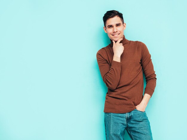 Portrait of handsome smiling model Sexy stylish man dressed in turtleneck sweater and jeans Fashion hipster male posing on grey background in studio Isolated Touching his chin