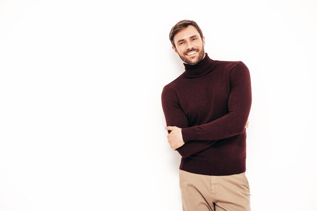 Portrait of handsome smiling model Sexy stylish man dressed in turtleneck sweater and trousers Fashion hipster male posing near white wall in studio Isolated