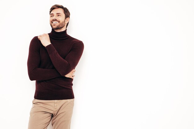 Portrait of handsome smiling model Sexy stylish man dressed in turtleneck sweater and trousers Fashion hipster male posing near white wall in studio Isolated