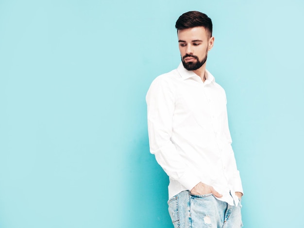 Portrait of handsome smiling model Sexy stylish man dressed in white shirt and jeans Fashion hipster male posing near blue wall in studio Isolated