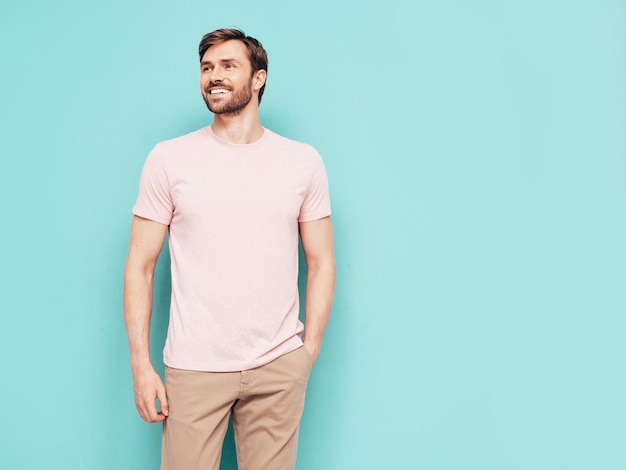 ハンサムな笑顔のスタイリッシュなヒップスターランバーセクシャルモデルの肖像ピンクのTシャツとズボンに身を包んだセクシーな男性スタジオの青い壁に分離されたファッションの男性