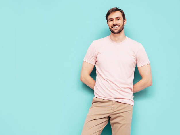 Portrait of handsome smiling stylish hipster lambersexual model Sexy man dressed in pink Tshirt and trousers Fashion male isolated on blue wall in studio