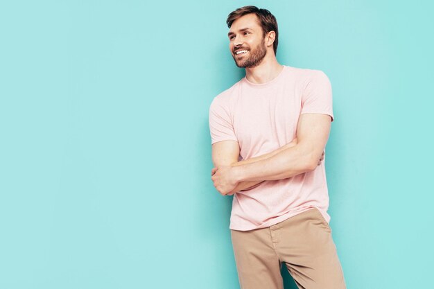 Portrait of handsome smiling stylish hipster lambersexual model Sexy man dressed in pink Tshirt and trousers Fashion male isolated on blue wall in studio