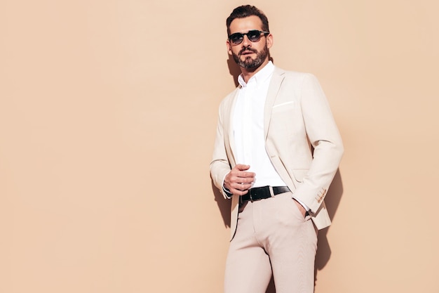 Free photo portrait of handsome smiling stylish hipster lambersexual model sexy modern man dressed in elegant white suit fashion male with curly hairstyle posing in studio near beige wall in sunglasses