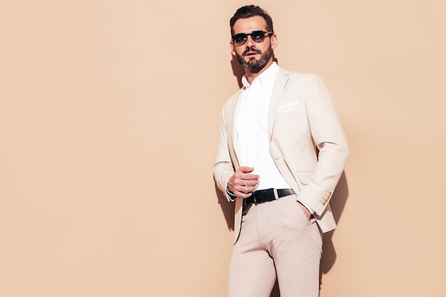 Portrait of handsome smiling stylish hipster lambersexual model Sexy modern man dressed in elegant white suit Fashion male with curly hairstyle posing in studio near beige wall In sunglasses