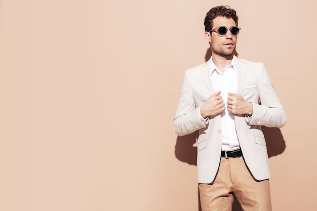 Portrait of handsome smiling stylish hipster lambersexual model Sexy modern man dressed in elegant white suit Fashion male with curly hairstyle posing in studio near beige wall In sunglasses