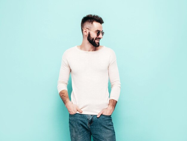흰색 스웨터와 청바지를 입은 잘생긴 웃고 있는 세련된 힙스터 램버섹슈얼 모델남자의 초상화 격리된 선글라스를 쓴 스튜디오의 파란색 벽 근처에서 포즈를 취하는 패션 남성