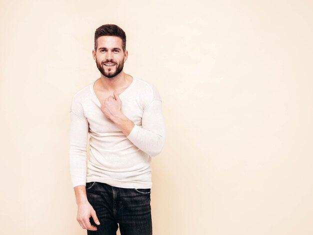 Portrait of handsome smiling stylish hipster lambersexual modelMan dressed in white sweater and jeans Fashion male posing near wall in studio