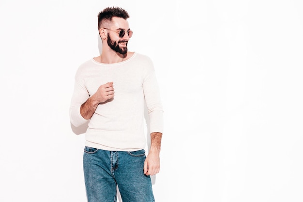 Portrait of handsome smiling stylish hipster lambersexual modelMan dressed in white sweater and jeans Fashion male posing near white wall in studio in sunglasses Isolated