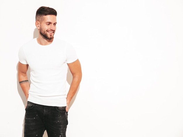 Portrait of handsome smiling stylish hipster lambersexual modelMan dressed in white tshirt and jeans Fashion male posing near wall in studio Isolated