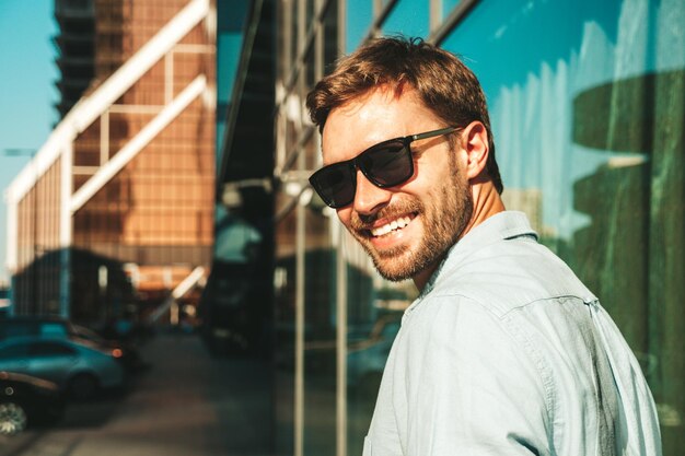 Portrait of handsome smiling stylish hipster lambersexual modelModern man dressed in blue shirt Fashion male posing in the street background near skyscrapers in sunglasses