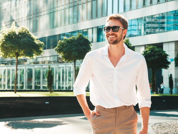Portrait of handsome smiling stylish hipster lambersexual modelModern man dressed in white shirt Fashion male posing in the street background near skyscrapers in sunglasses Outdoors at sunset