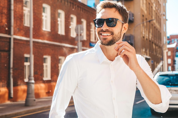 Portrait of handsome smiling stylish hipster lambersexual modelModern man dressed in white shirt Fashion male posing on the street background in sunglasses Outdoors at sunset