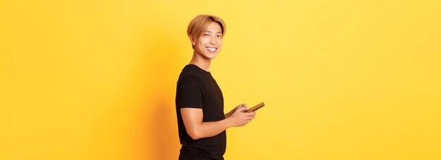 Portrait of handsome stylish asian guy in black outfit using mobile phone and turning head at camera