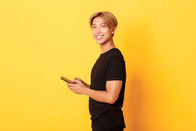 Portrait of handsome stylish asian guy in black outfit, using mobile phone and turning head with satisfied smile, yellow wall