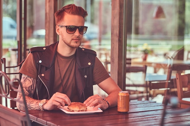 Portrait of a handsome stylish hipster guy sits at a table, ordered a hamburger and soda, dines at a roadside cafe