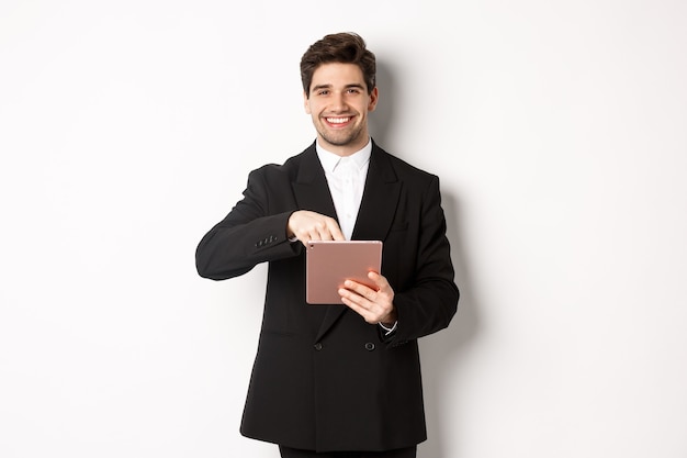 Portrait of handsome, stylish male entrepreneur in black suit pointing at digital tablet, showing something online, standing against white background