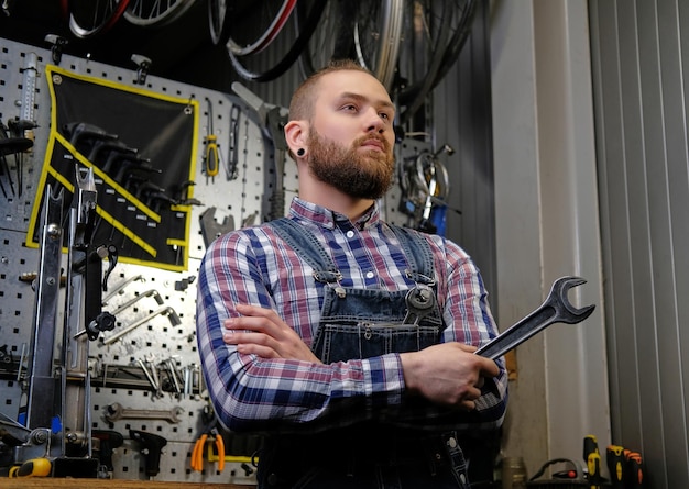 Free photo portrait of a handsome stylish male with beard and haircut wearing a flannel shirt and jeans coverall, holds steel wrench, standing in a workshop against wall tools.