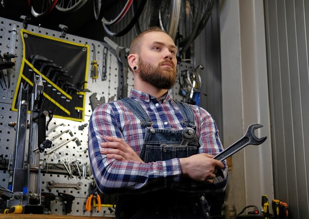 Portrait of a handsome stylish male with beard and haircut wearing a flannel shirt and jeans coverall, holds steel wrench, standing in a workshop against wall tools.