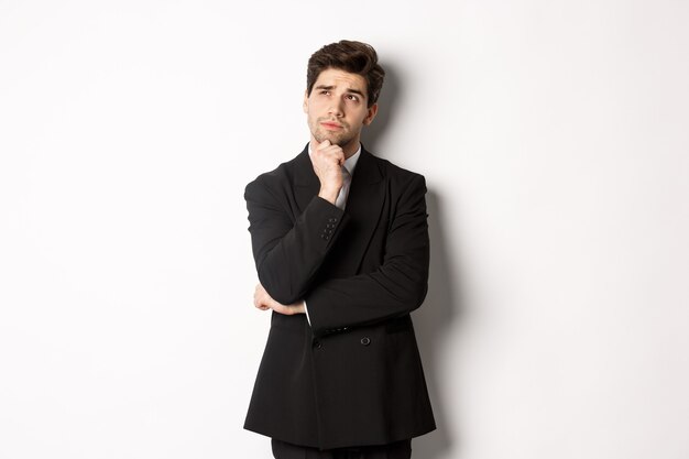 Portrait of handsome and stylish man in suit, thinking and looking at upper left corner, imaging something about christmas holidays, standing over white background