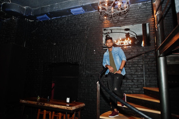 Portrait of handsome successful bearded south asian young indian freelancer in blue jeans shirt standing in night club on stairs