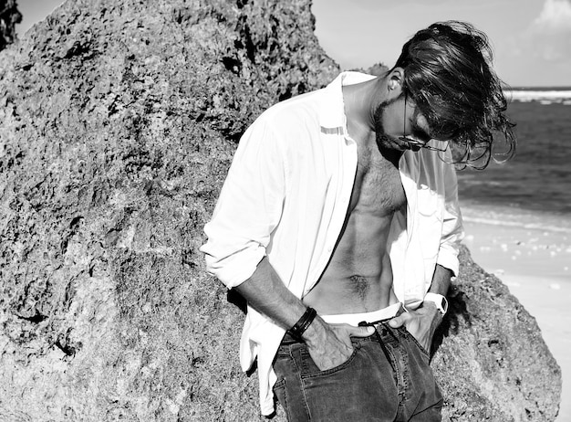 Portrait of handsome sunbathed fashion man model wearing white shirt clothes in glasses posing near rocks on summer beach on blue sky 