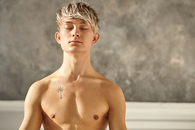 Портрет красивого татуированного мужчины, практикующего йогу в помещении, закрывая глаза во время медитации, имея мирный взгляд, концентрируясь на своем дыхании. Молодой учитель-мужчина без рубашки делает медитацию в тренажерном зале
