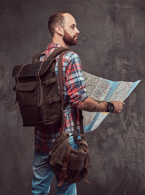 Portrait of a handsome tattooed traveler in a flannel shirt with a backpack, holds a map, standing in a studio. Isolated on a gray background.