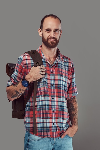 Portrait of a handsome tattooed traveler in a flannel shirt with a backpack, standing in a studio. Isolated on a gray background.