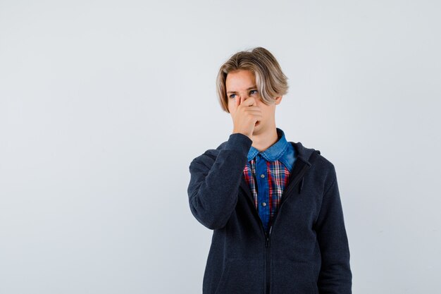 Portrait of handsome teen boy pinching nose, smelling something disgusting in shirt, hoodie and looking displeased front view