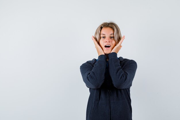 Portrait of handsome teen boy with hands on cheeks in hoodie and looking excited front view