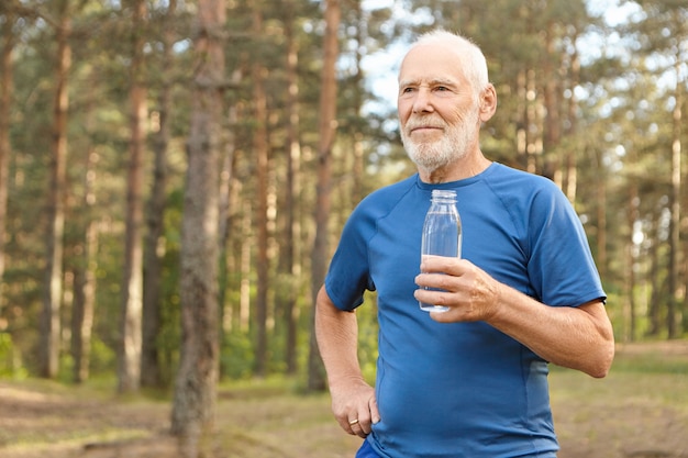 Portrait of handsome tired European senior elderly male in t-shirt holding glass bottle, enjoying fresh drinking water after running exercise in forest, catching his breath, looking around