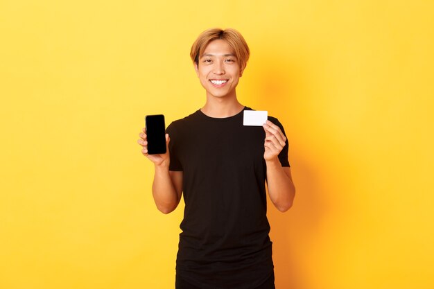 Portrait of handsome young asian guy showing smartphone screen, banking app and credit card, standing yellow wall and smiling.