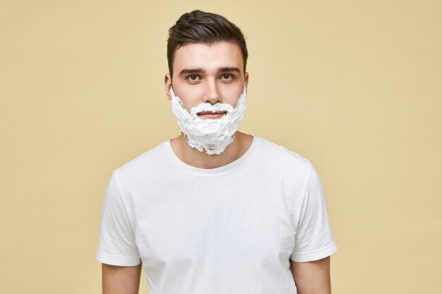 ハンサムな若い白人ブルネットの男性の肖像画は、彼の顔に適用された白いひげそりの泡で隔離され、剃毛のために肌を準備しています。朝のルーチン、男らしさ、美容とケアのコンセプト
